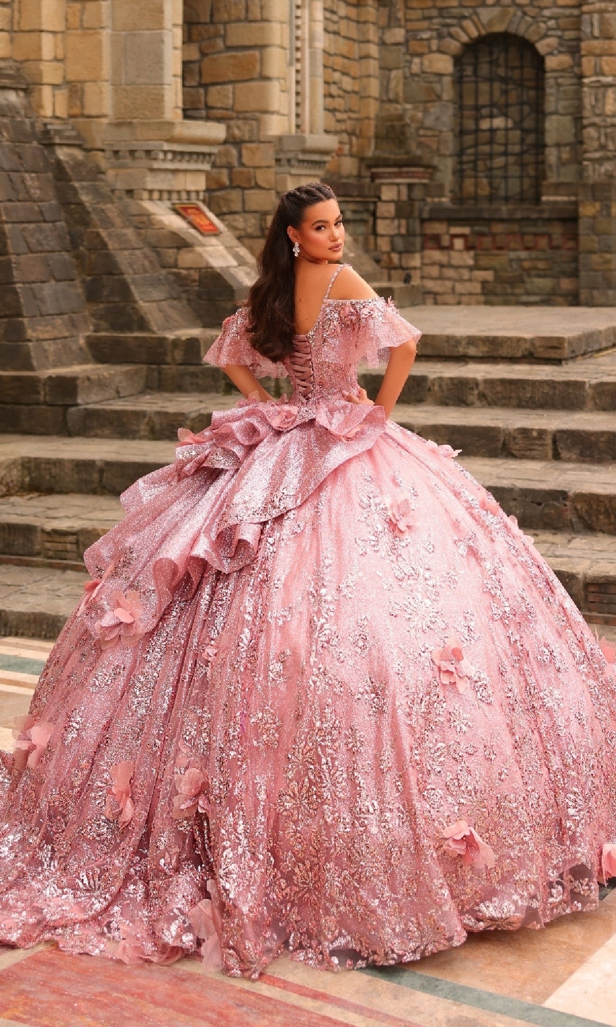 A young woman stands on stone steps in the Amarra 54229 Glitter Tulle Quinceañera Dress, its beading and ornate pink floral details sparkling against the backdrop of stone walls and windows.